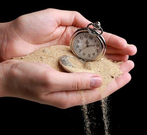 Hands holding a pocket watch with sand pouring through the fingers. The sand flows downward, partially covering the watch, symbolizing the passage of time.