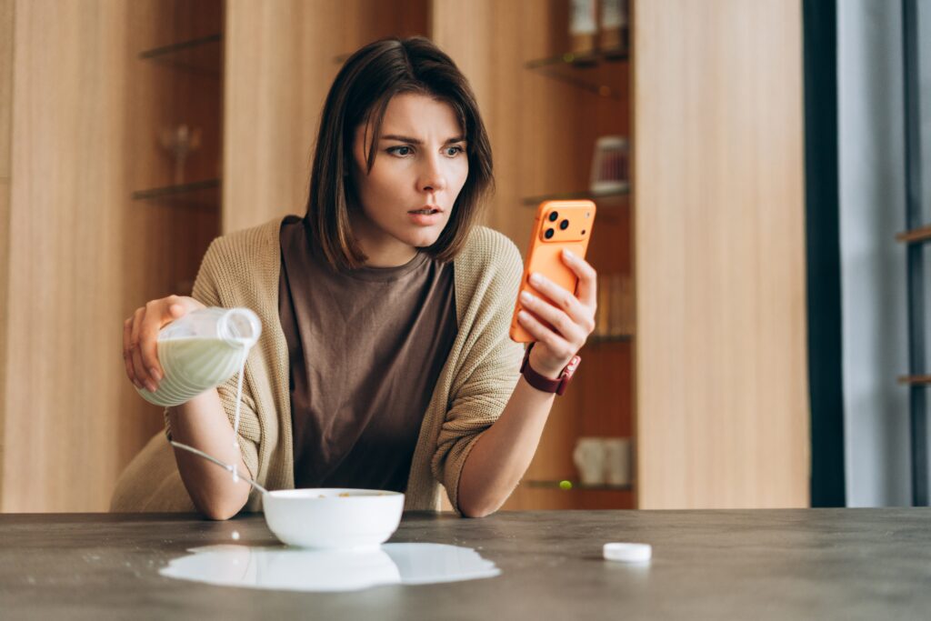 A woman mistakenly missing a bowl while pouring milk. Her eyes wide open with a look of shock on her face while looking at her phone in the other hand.